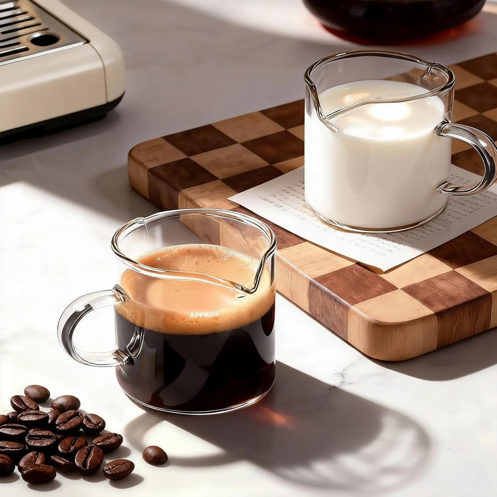 Glass coffee cup with espresso, milk pitcher, and coffee beans on a checkered surface.