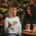 Two women sitting at a table with pastries and drinks in a floral setting, one wearing a light gray sweatshirt with a croissant illustration.