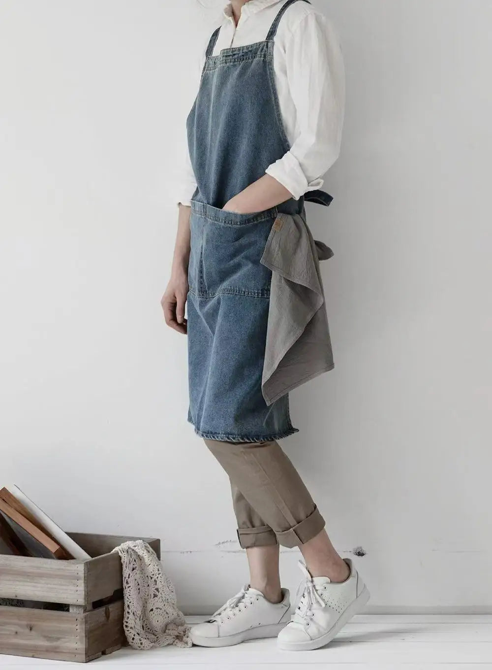 Person wearing a denim apron over a white shirt, standing against a plain wall with a wooden crate and fabric in the foreground.