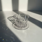 A clear ribbed glass coffee dripper resting on a matching round glass coaster, set on a light gray textured surface with strong sunlight and shadows.