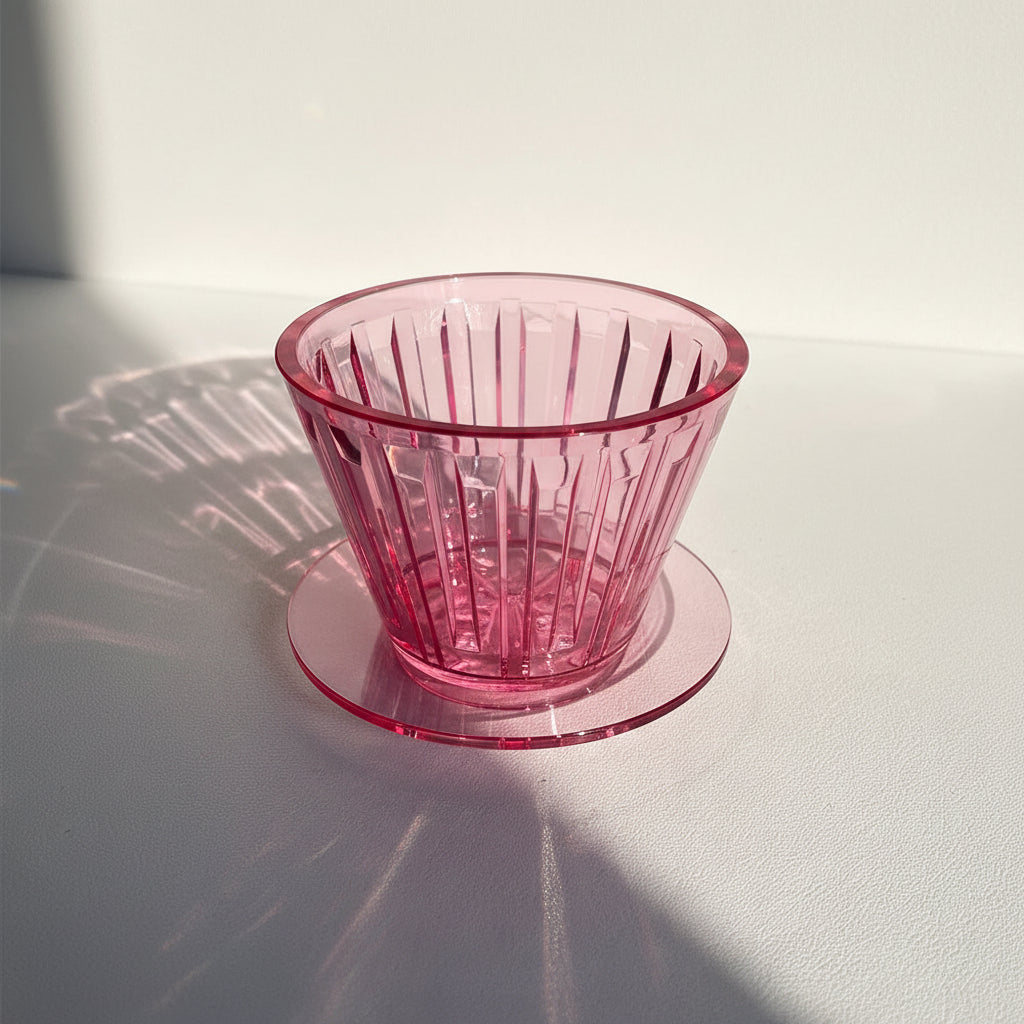 A translucent pink glass cup with a vertical ribbed pattern sits on a matching round coaster on a light-colored surface, captured in bright natural sunlight with a strong shadow.
