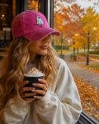A woman sitting by a window in a cafe, wearing a pink baseball cap with a white ghost graphic and holding a whipped coffee drink. The background shows an autumn park scene with yellow and orange leaves.