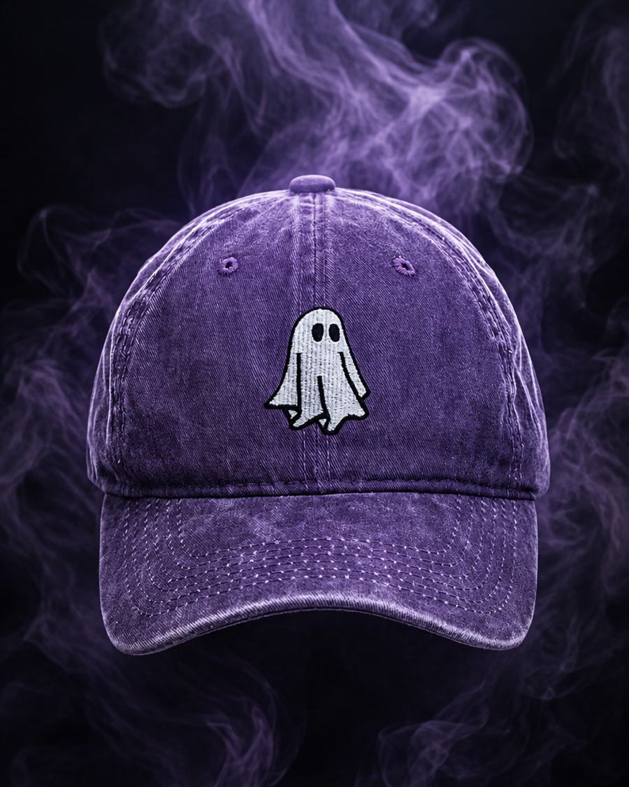 A purple washed baseball cap with a white embroidered ghost patch on the front, set against a dark background with purple smoke.