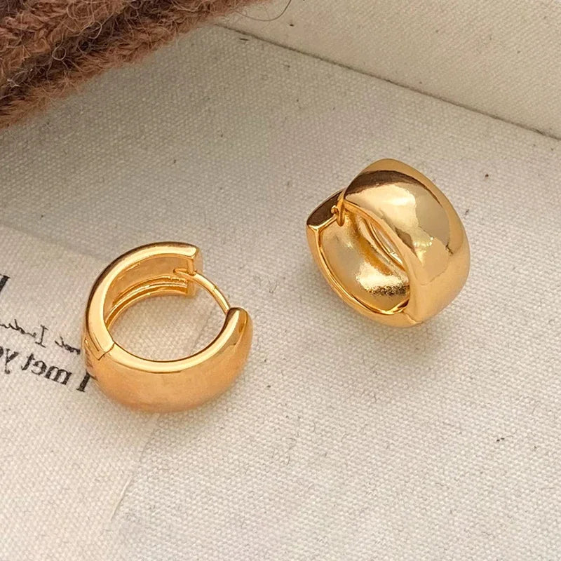 A pair of chunky, polished gold huggie hoop earrings displayed on a textured beige fabric surface.