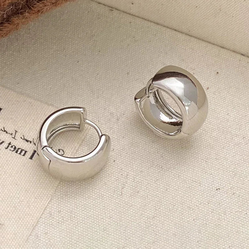 A pair of polished silver huggie hoop earrings resting on a textured beige fabric surface.