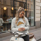 Woman wearing a light gray Le Croissant Crewneck holding a coffee cup on a scooter in an urban setting