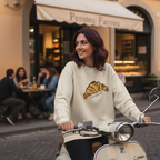 A woman wearing a Le Croissant Crewneck riding a scooter in an urban setting with a café in the background