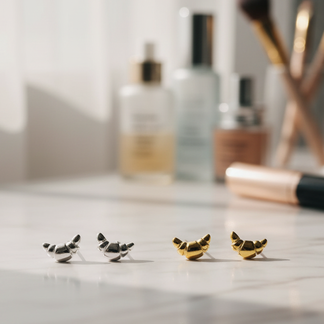 Mini croissant silver & gold earrings on a marble surface with blurred makeup items in the background. 