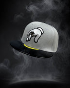 A gray snapback cap with a black flat brim, featuring a white and black graphic of a hand on the front and a yellow accent line above the brim, set against a dark smoky background.