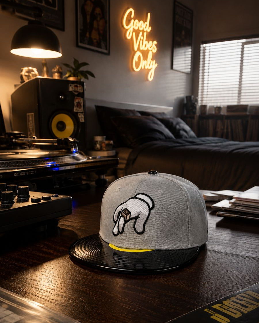 A gray snapback cap with a black brim and a white graphic of a hand holding a microphone, resting on a wooden table in a dimly lit bedroom setting.
