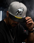 A man wearing a gray and black snapback cap with a white and black embroidered graphic on the front, holding the brim. The background is dark with smoke and DJ equipment.