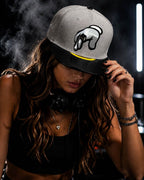 A woman DJ wearing a gray and black baseball cap with a hand logo, a black crop top, and headphones around her neck, standing behind a turntable with smoke in the background.