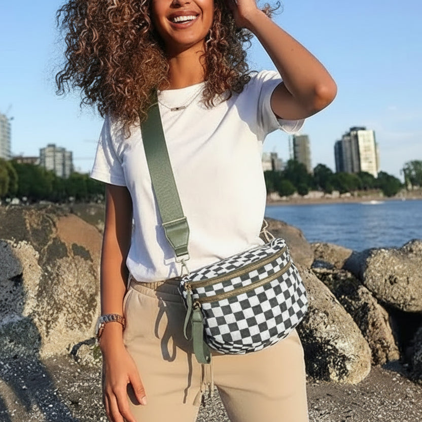 Woman wearing an olive + white check Baye Bag standing on a rocky beach with a cityscape in the background.