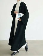 Person wearing a long black coat over a white turtleneck, holding a book against a plain background