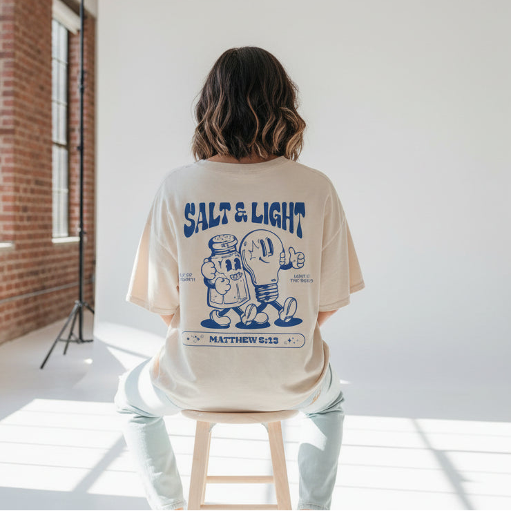 Female model wearing a 'Salt & Light' t-shirt sitting on a stool in a studio setting.