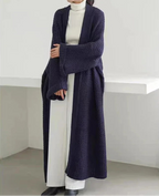 Person wearing a long dark blue knitted cardigan over a white outfit in a minimalistic setting.
