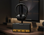 A black and gold audio amplifier with a stand holding black over-ear headphones, set on a wooden desk in a dimly lit home office environment with a computer monitor in the background.