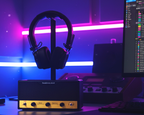 Black over-ear headphones displayed on a stand atop a small black and gold audio amplifier on a desk, with a computer monitor showing audio software in the background under purple and blue neon lighting.