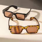 Two pairs of sunglasses, one with black frame and brown lenses, the other with tortoiseshell frame and brown lenses, on a light surface.