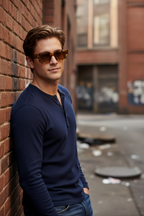 A man wearing brown-tinted sunglasses and a navy blue long-sleeve henley shirt leans against a red brick wall in an urban alleyway.