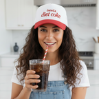 Woman wearing a 'I need a Diet Coke' cap holding a glass of soda in a kitchen.